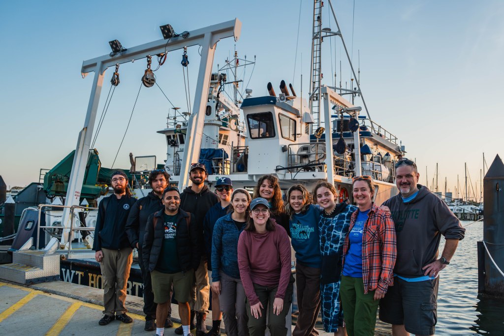 A group of twelve people in front of a ship. The water is in the background and the sky is hazy.